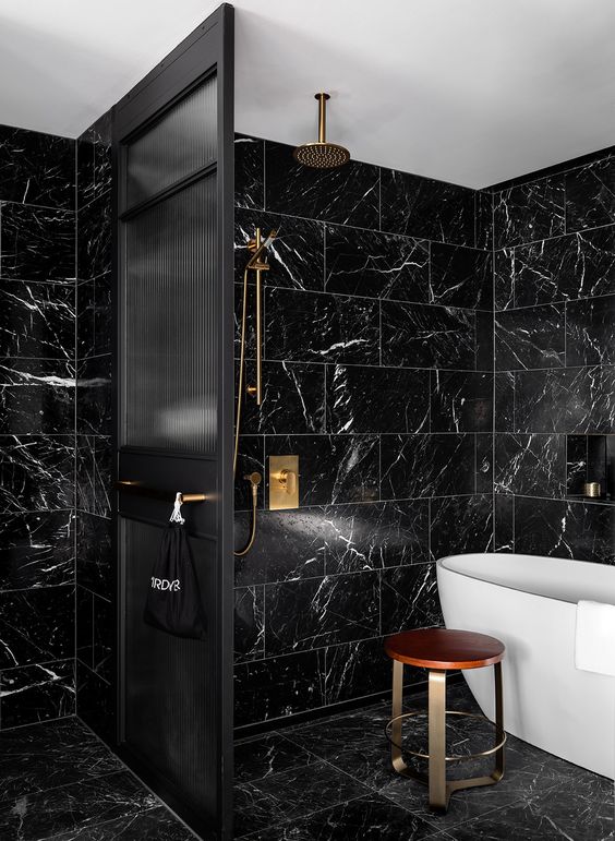 a stylish black bathroom clad with marble tiles, with a white tub for a contrast and brass touches