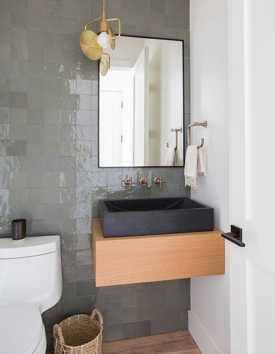 a contemporary bathroom with a shiny grene tile wall, a wooden vanity, a black square stone sink and a black framed mirror
