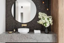 a gorgeous modern bathroom with a grey tile wall, black lamps, a grey stone vanity and a round mirror