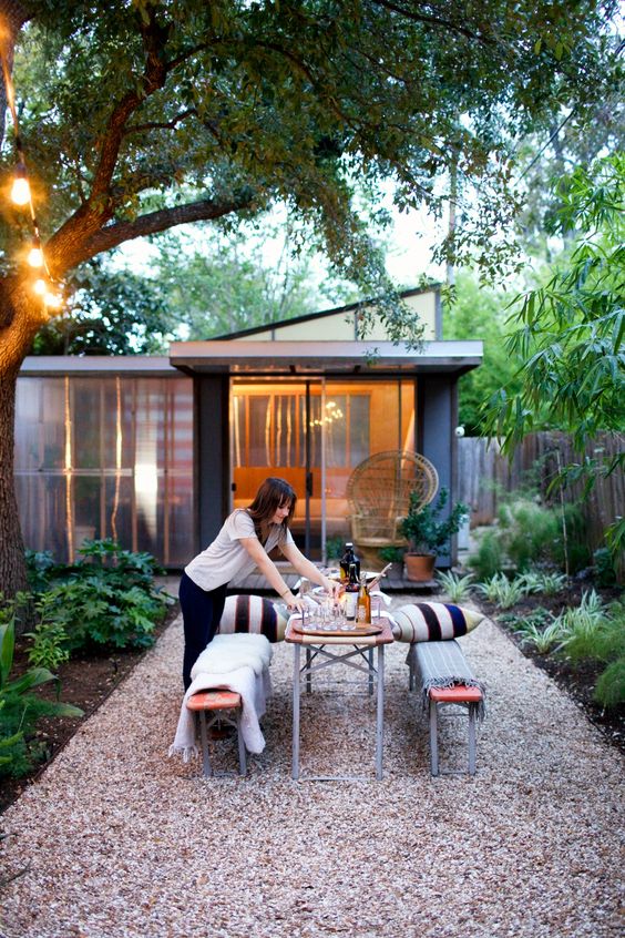 a modern backyard with greenery and trees, wooden and metal benches and a dining table plus striped pillows