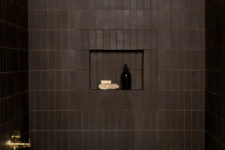 a refined moody bathroom clad with chocolate brown tiles, with chic gold fixtures, a white tub and a niche for storage