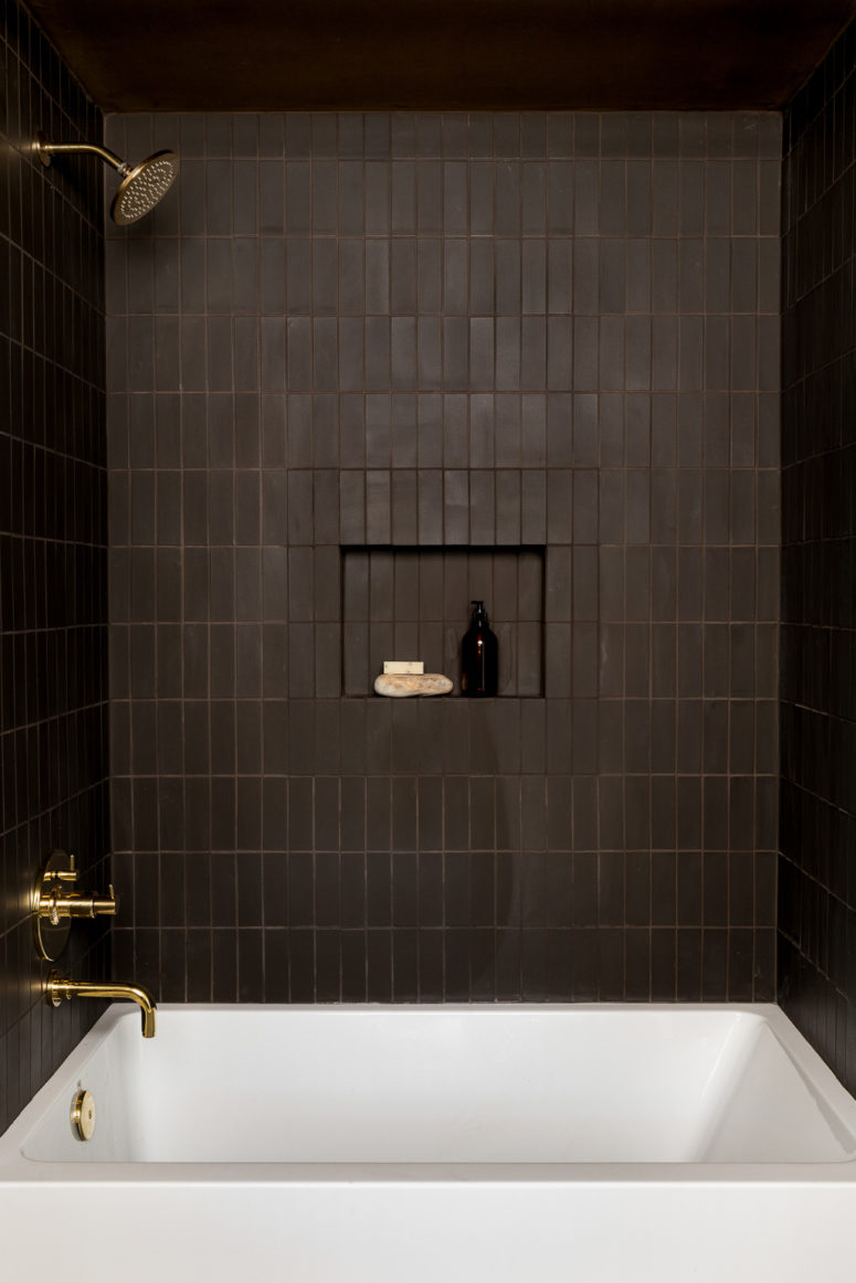 a refined moody bathroom clad with chocolate brown tiles, with chic gold fixtures, a white tub and a niche for storage