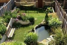 a small and cool backyard turned into a garden, with a pond, a green lawn, some blooms and plants and a bench