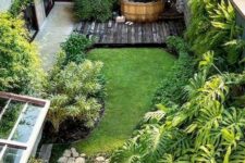 a small and cool backyard with a green lawn, greenery around, some rocks and a deck with a hot tub in the corner