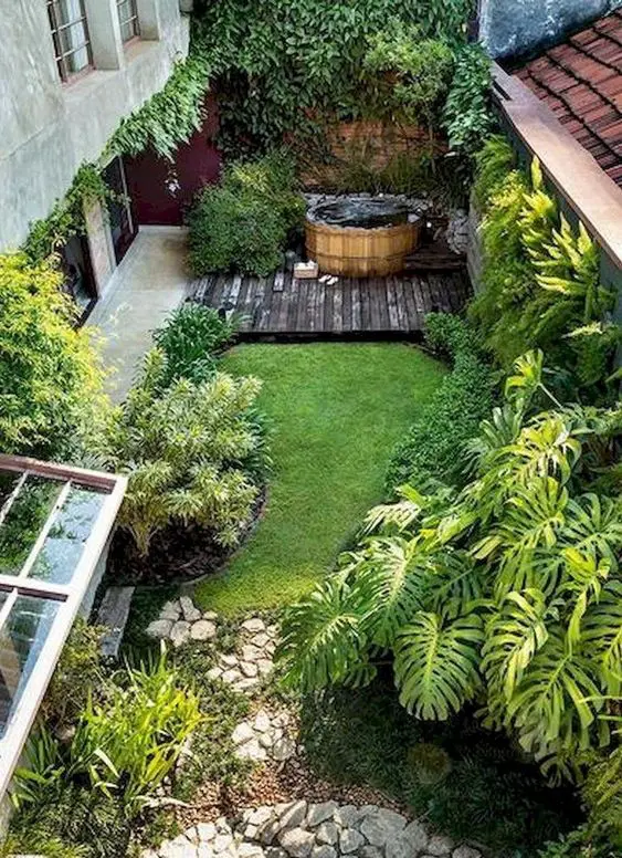 a small and cool backyard with a green lawn, greenery around, some rocks and a deck with a hot tub in the corner