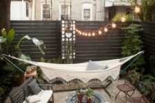 a small backyard with a couple of chairs, a bench and a coffee table, a hammock, some greenery and string lights over the space