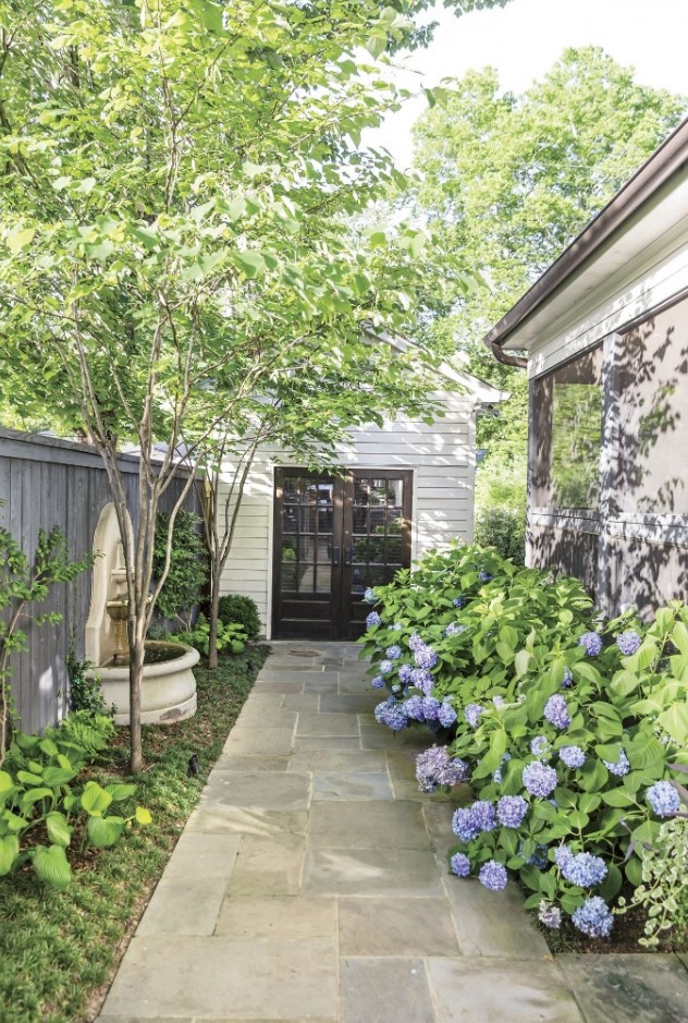 a small backyard with greenery and a small fountain, with lush florals lining up the wall of the house is a dreamy space