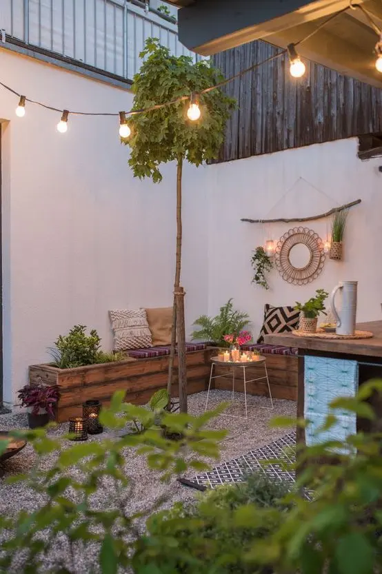 a small boho backyard with gravel on the ground, a wooden corner bench, some greenery, lights, boho pillows and lights