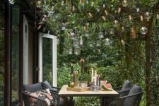 a small terrace with a wooden deci, a table and black chairs, beams with greenery and lights over the space