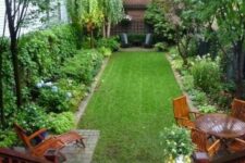 a small yet lovely backyard with a green lawn, some greenery and blooms around, a wooden dining set and a lounger and a wooden deck