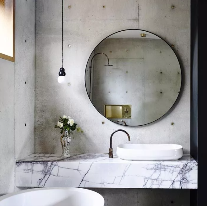 an industrial bathroom with concrete walls, a marble slab vanity, an oversized round mirror and a pendant lamp