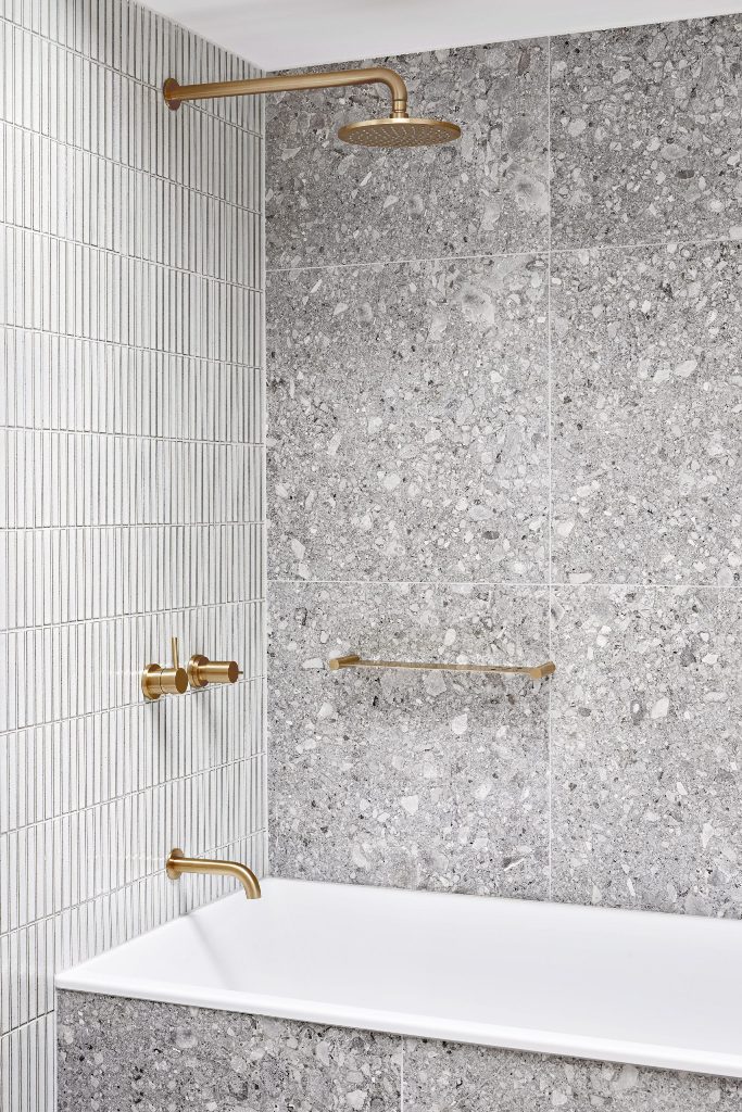 The bathroom is clad with grey stone and white skinny tiles and is accented with gold fixtures