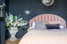 a chic moody bedroom with black walls and a chest for storage, a light pink bed, refined lighting and blooms