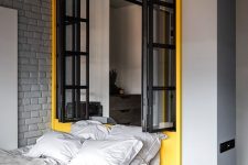 a modern industrial bedroom with light grey walls, a grey brick wall, a yellow zone with a window and a yellow bed with grey bedding