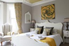 a refined bedroom with dove grey walls, a pale yellow ceiling, elegant creamy furniture, a crystal chandelier and touches of mustard