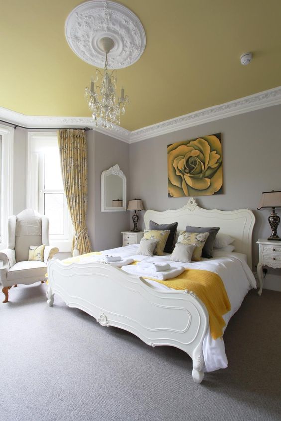 a refined bedroom with dove grey walls, a pale yellow ceiling, elegant creamy furniture, a crystal chandelier and touches of mustard