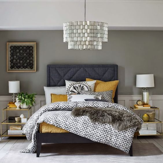 a shiny and elegant bedroom with grey walls, white paneling, a graphite grey bed, a shiny chandelier, gold frame nightstands and mustard bedding