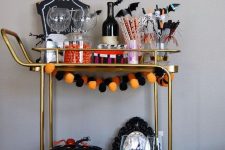 a bold Halloween bar cart with a bright pompom garland, a black spiked pumpkin, skulls, bats on the wall