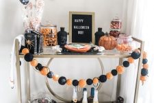 a bright Halloween bar cart with an orange and black garland, a sign, bats on the wall, pumpkins, candies and a gnome