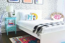 a colorful kid room with a Dolmatin wall, colorful bedding and a rug, bold artworks, a blue nightstand and a rattan chair