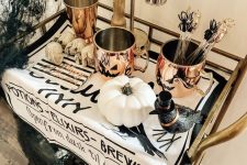 a cool Halloween bar cart with copper mugs, a crow, white pumpkins, skulls, black spiderwebs and shiny touches