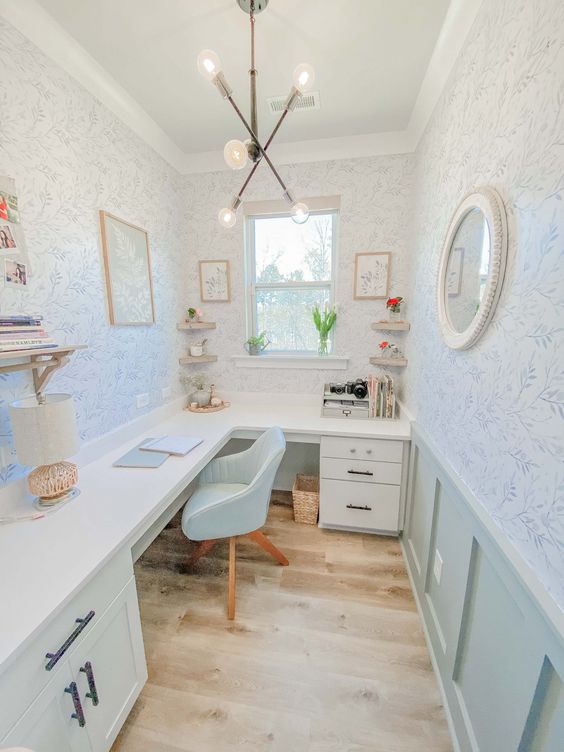 A cottage home office with printed wallpaper, mint paneling, an L shaped desk, some decor and open shelves