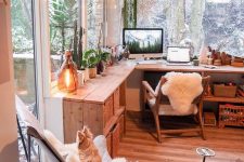 a cozy boho working nook with a wooden L-shaped desk, a stained chair, a butterfly one, some potted plants and decor