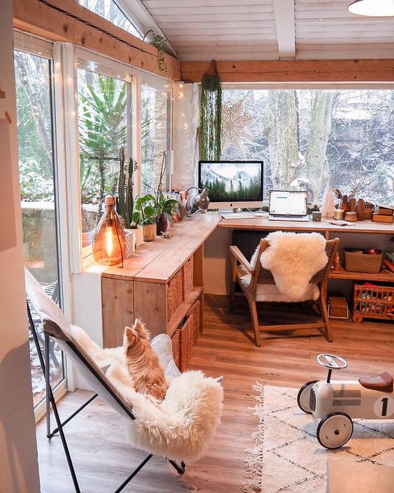 A cozy boho working nook with a wooden L shaped desk, a stained chair, a butterfly one, some potted plants and decor