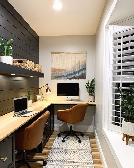 A farmhouse home office with a black planked wall, an L shaped desk, a couple of leather chairs, potted plants and a macrame piece