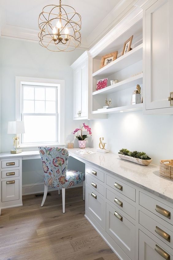 A glam farmhouse home office with white cabinetry, a large L shaped desk, a printed chair, some decor and a cool gold chandelier