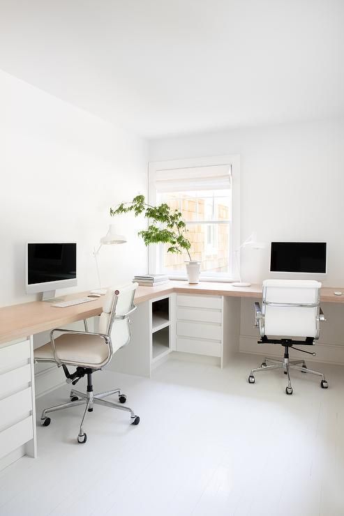 A minimalist white home office with an L shaped desk, white chairs, storage units with drawers and potted greenery