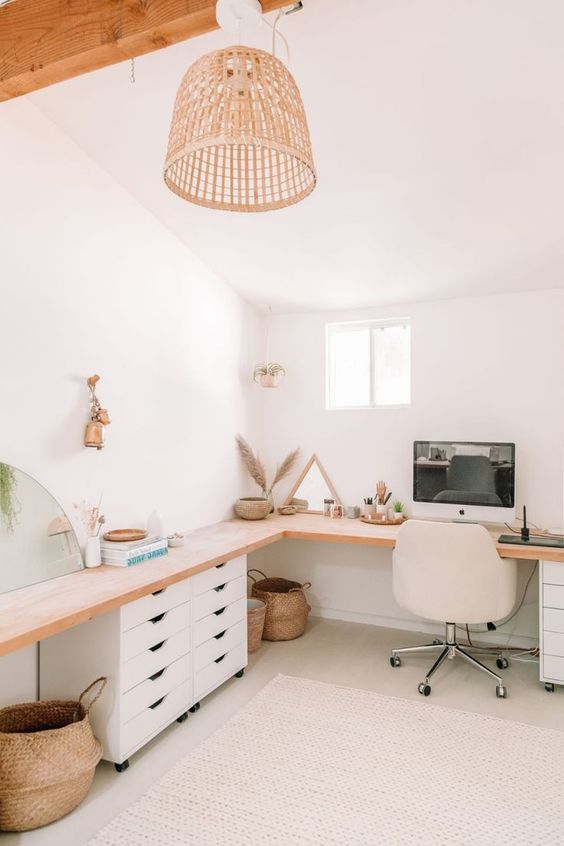 A neutral boho home office with a large L shaped desk with some drawers, a white chair, some decor and baskets and a pendant lamp
