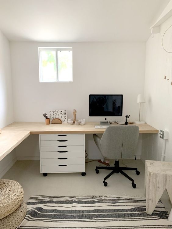 A neutral farmhouse home office with an L shaped desk, drawers, a grey chair, a stool, jute poufs, some decor and a lamp