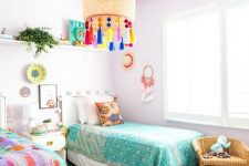 a pastel shared kids’ bedroom with lavender walls and a pouf, colorful bedding and art and a lamp with colorful tassels
