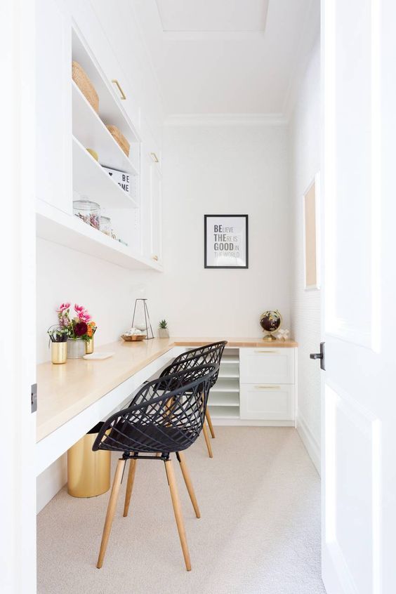A small white home office with an L shaped desk with storage, black chairs, a storage unt, some decor and blooms