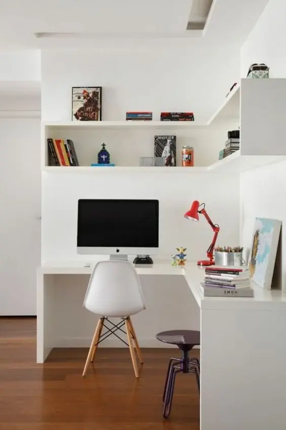 a small white working nook with a corner desk and a box corner shelf, a chair and a stool, a red table lamp and some decor