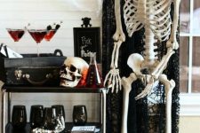 a spooky Halloween bar cart with black glasses, a black chest with drinks, a skull and skeletons around