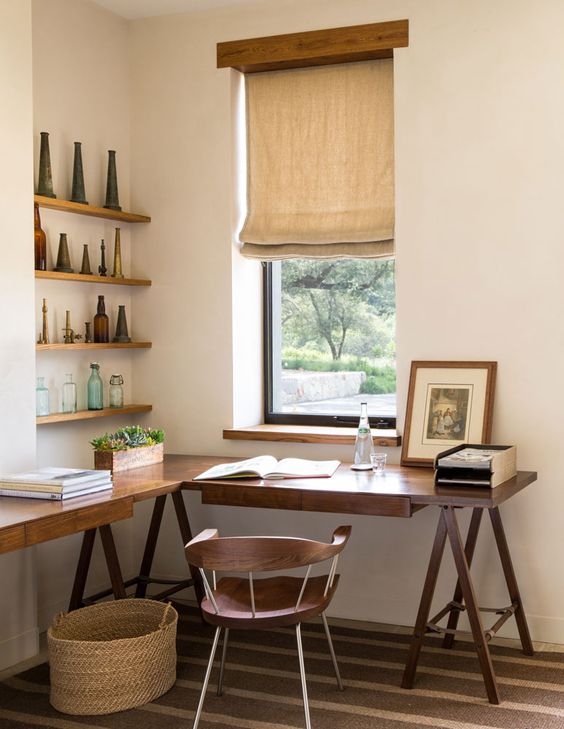 A stylish work nook with an L shaped trestle desk, a stained chair, open shelves and decor, a burlap curtain