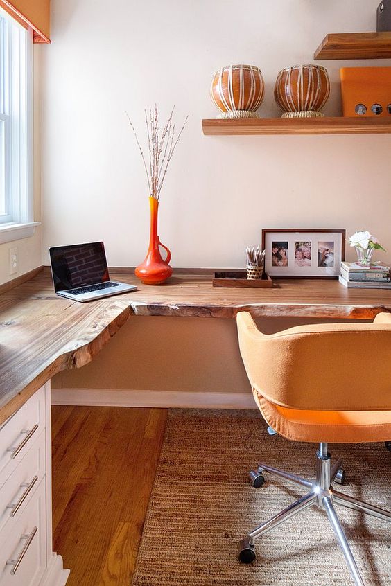 A warm colored home office with a live edge corner desk, a yellow chair, open shelves and an orange vase