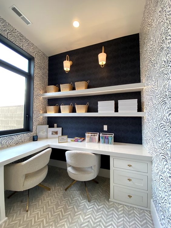 A whimsical home office with a black wall and printed wallpaper, a white L shaped desk, white chairs, open shelves and some decor