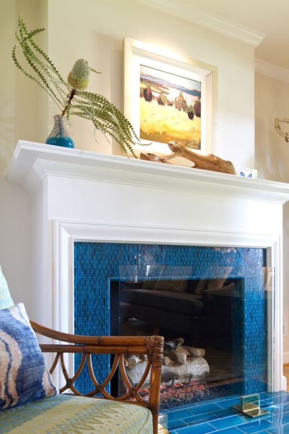 a fireplace surrounded with bright blue tiles, with an elegant white mantel and some coastal art on it is ideal for a seaside home