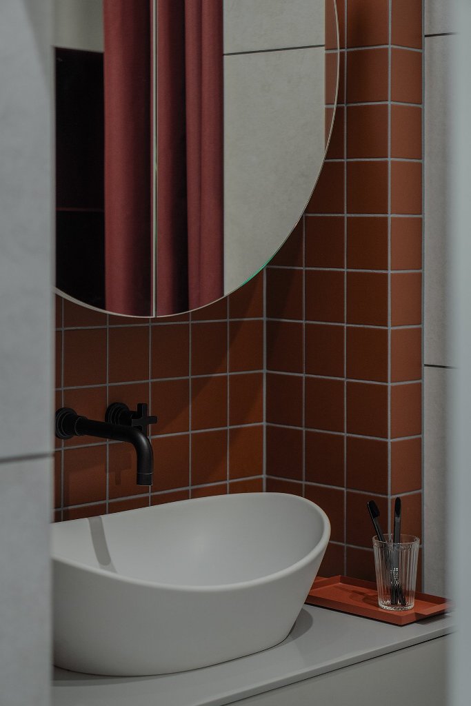 The bathroom is done with rust colored tiles, a round mirror and a vessel sink