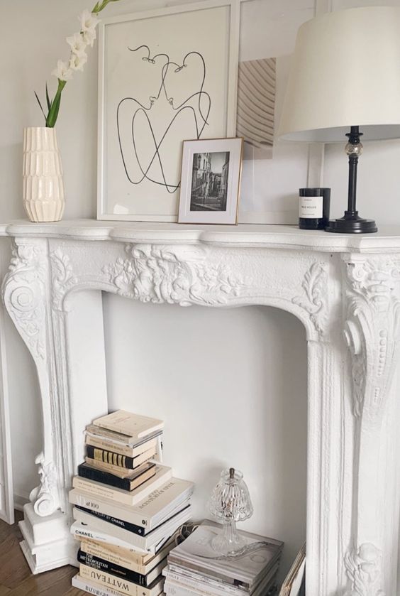 a French fireplace with books and a lamp, some art, a vase with blooms and a table lamp on the mantel