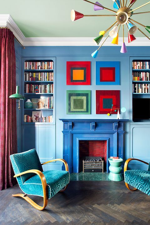 A blue living room with built in bookcases, a bright fireplace, teal printed chairs, bold artwork and a colorful chandelier