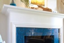 a fireplace surrounded with bright blue tiles, with an elegant white mantel and some coastal art on it