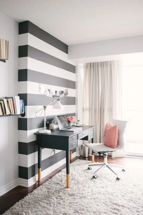 A light filled home office with a black and white striped accent wall, a black desk, a white chair, neutral textiles and pink touches is very chic
