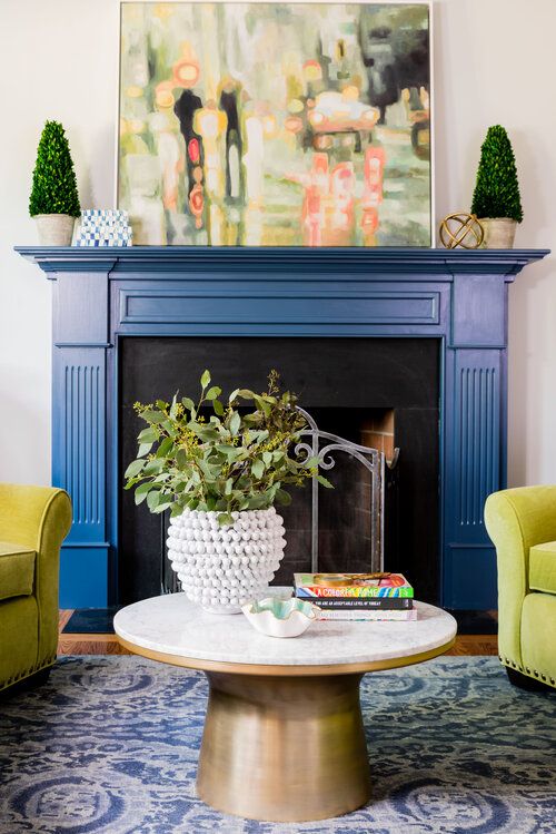 a living room with a fireplace and a blue mantel, potted plants and an artwork, lemon yellow chairs and a printed rug