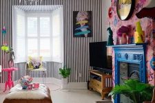 a maximalist living room with a striped wall, a bold floral one, a bright blue mantel over a vintage fireplace, a crystal chandelier and touches of hot pink