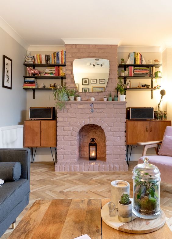 A mid century modern living room with cabinets, open shelves, a mauve brick fireplac,e a grey sofa, a pink chair and a coffee table