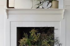 a non-working fireplace with a bucket with evergreens, an arrangement of mirrors and greenery on the mantel